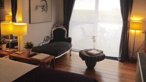 Habitación de hotel con una cama grande, un sillón oscuro, una mesita redonda de madera con un libro y un vaso, y dos lámparas a cada lado de la ventana.