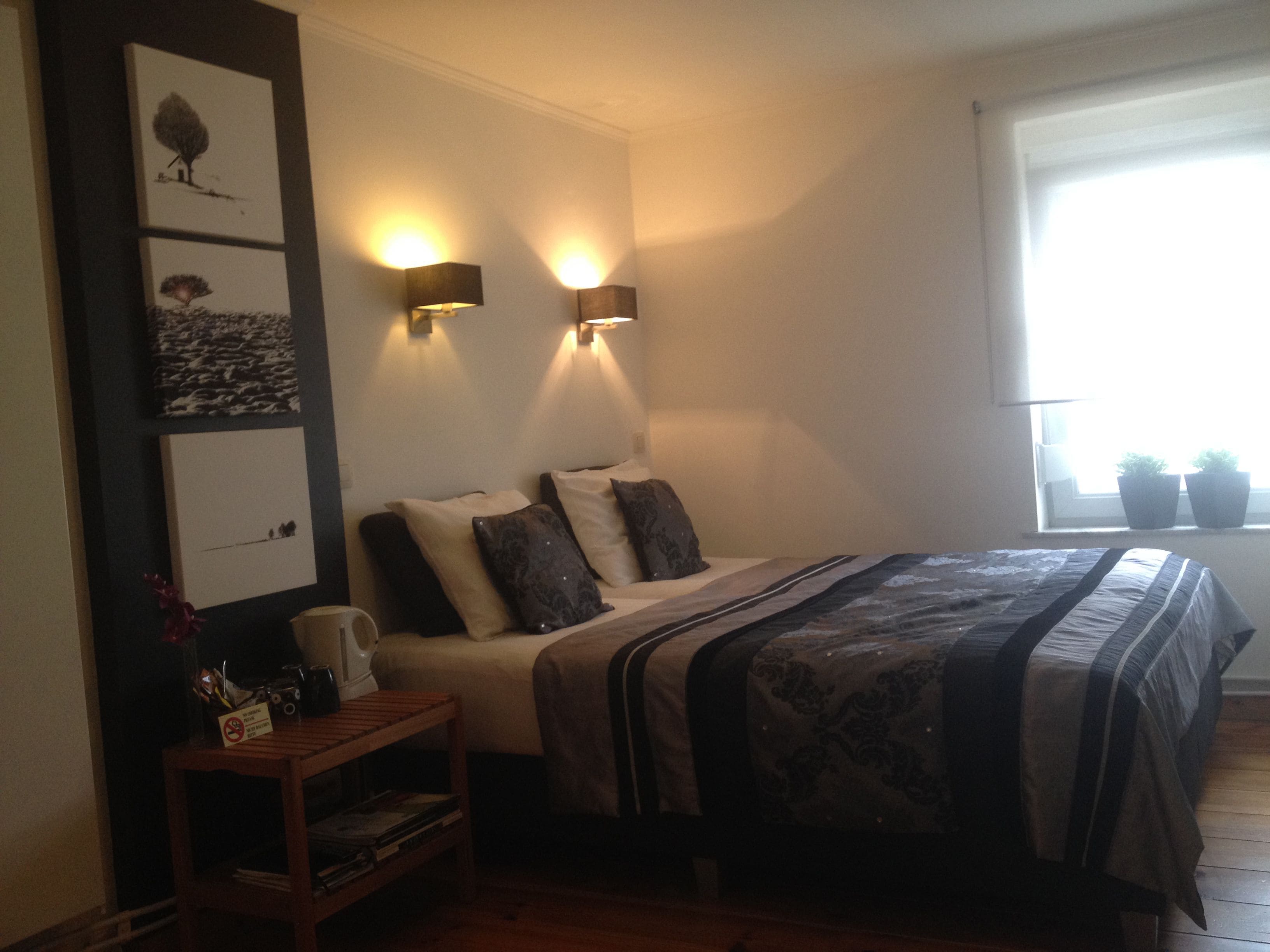 Une chambre à coucher avec un lit, deux lampes fixées au mur, une table de chevet et une fenêtre agrémentée de plantes en pot.