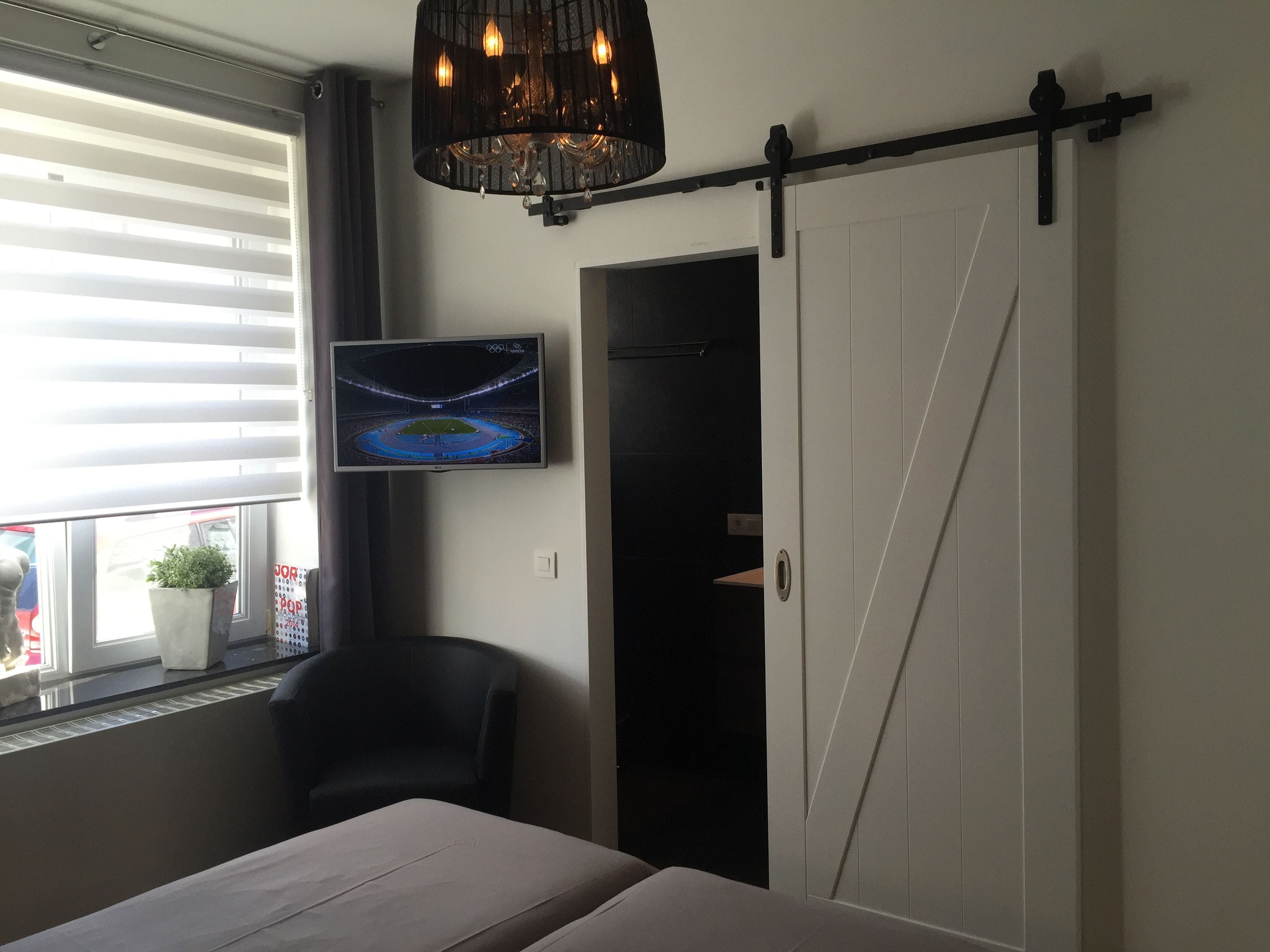 A bedroom with a bed, a TV mounted on the wall, a sliding door, and a window with blinds.