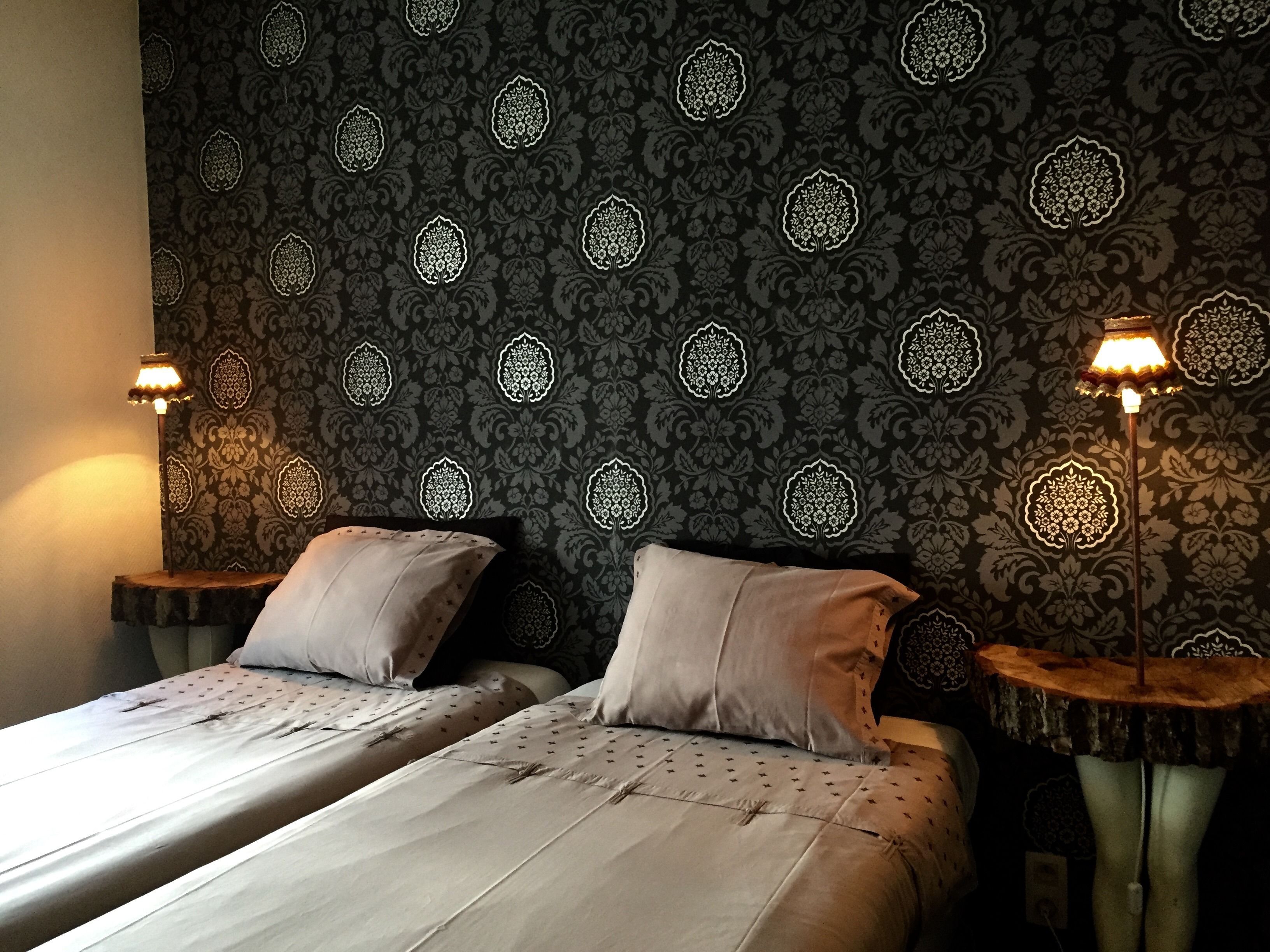 Ein Zimmer mit zwei Betten, jeweils mit Kissen und einem Nachttisch. Die Wände sind mit einem gemusterten Wandpapier tapeziert. An beiden Seiten der Betten befinden sich zwei Wandlampen.