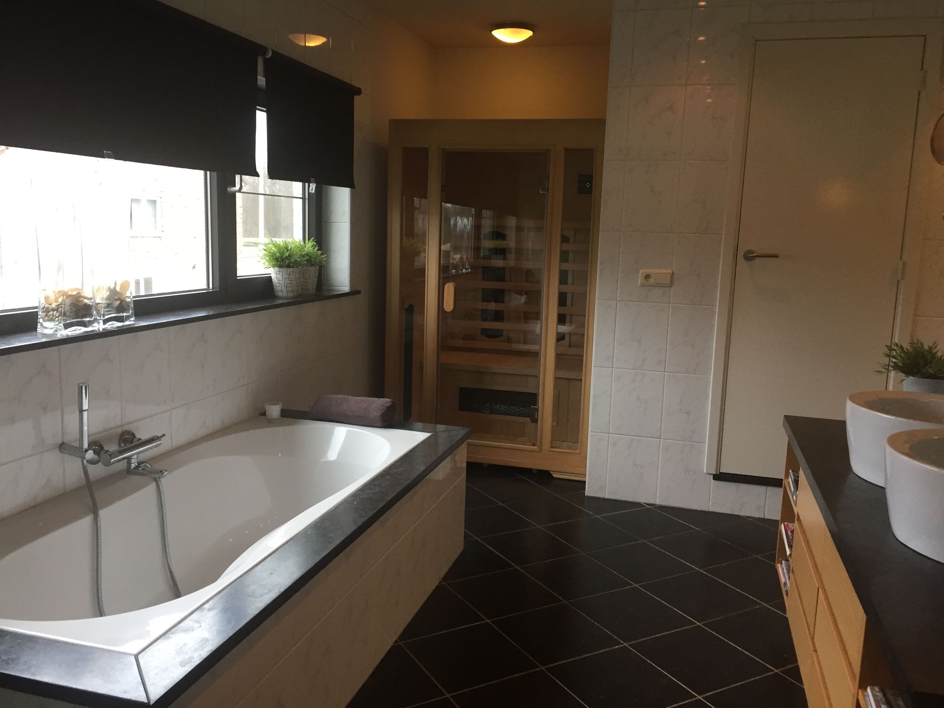 A modern bathroom with a large bathtub, a window with plants, and a wooden cabinet with books.