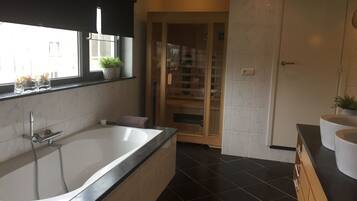 A modern bathroom with a large bathtub, a window with plants, and a wooden cabinet with books.