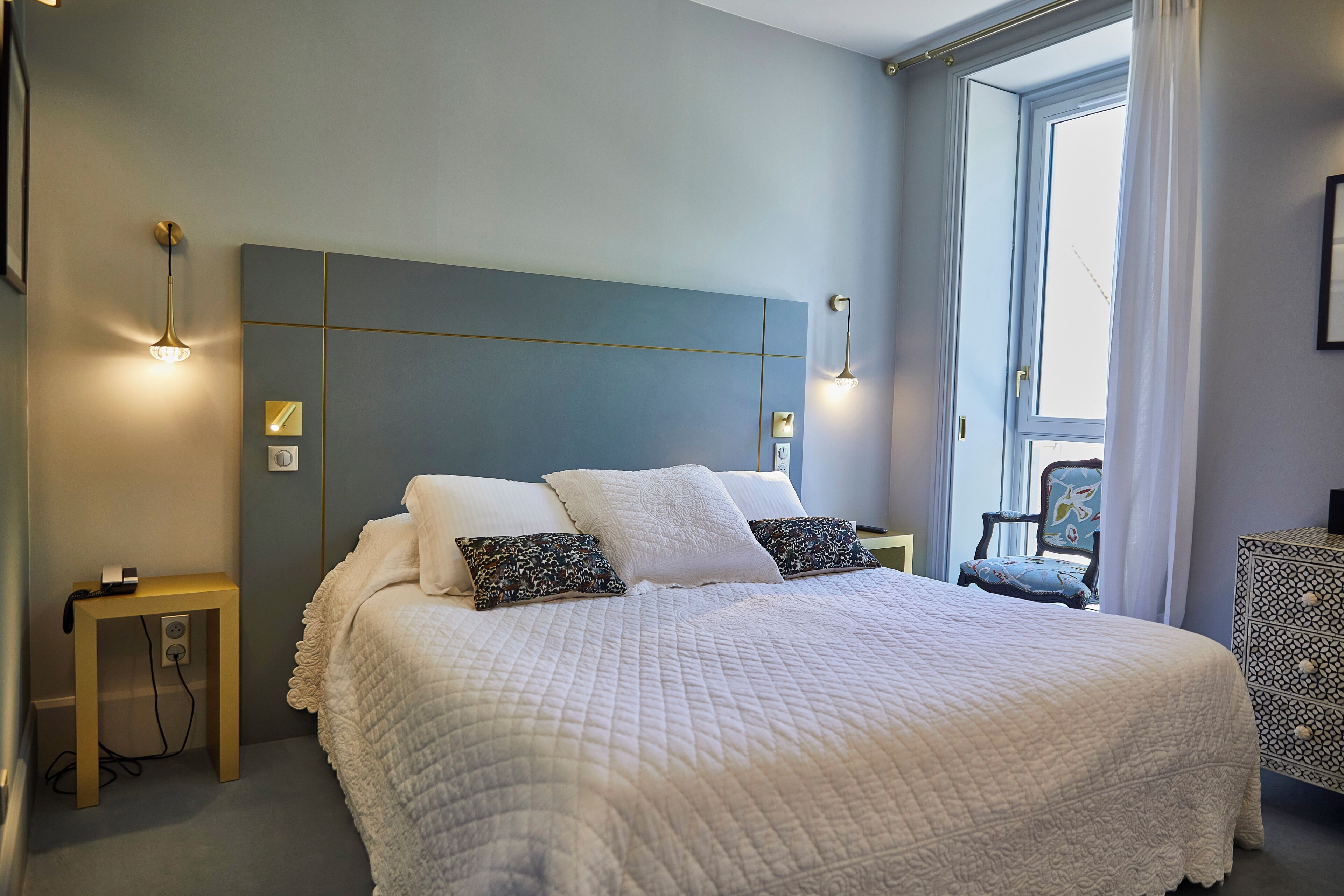 A neatly made bed with a quilted white cover, two pillows, bedside tables with lamps, and a window with curtains.