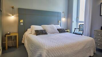 A neatly made bed with a quilted white cover, two pillows, bedside tables with lamps, and a window with curtains.