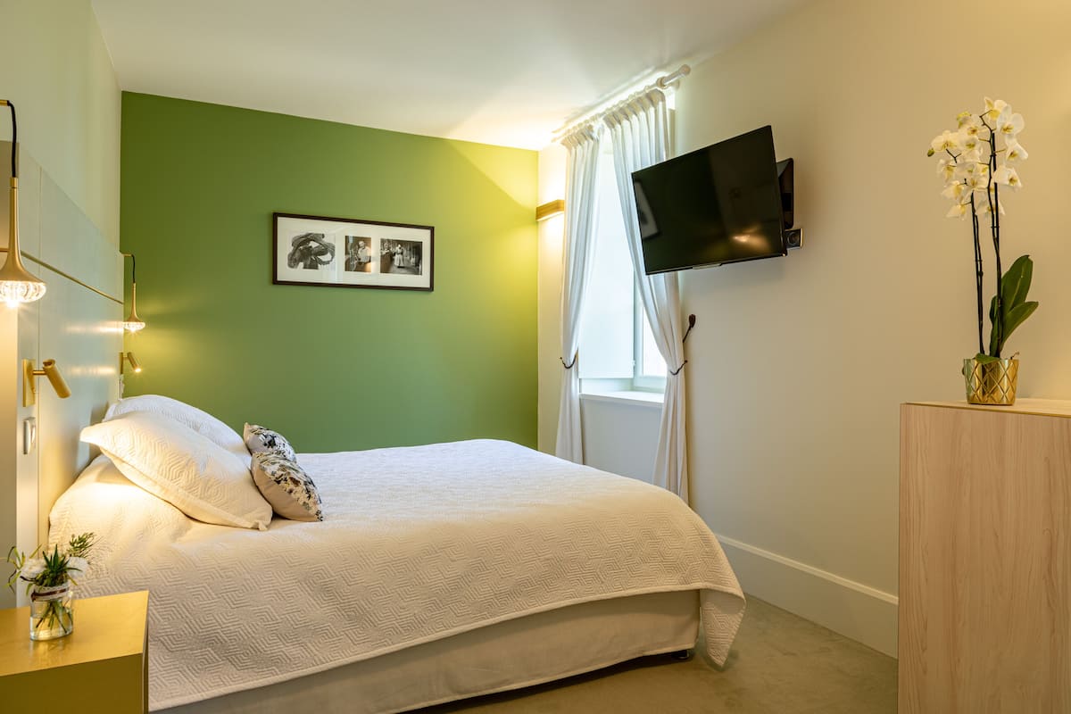 Ein Schlafzimmer mit einem Bett, einem flachbildfernseher an der Wand, einem Fenster mit Vorhängen und einer kleinen Pflanze auf einem Nachttisch.