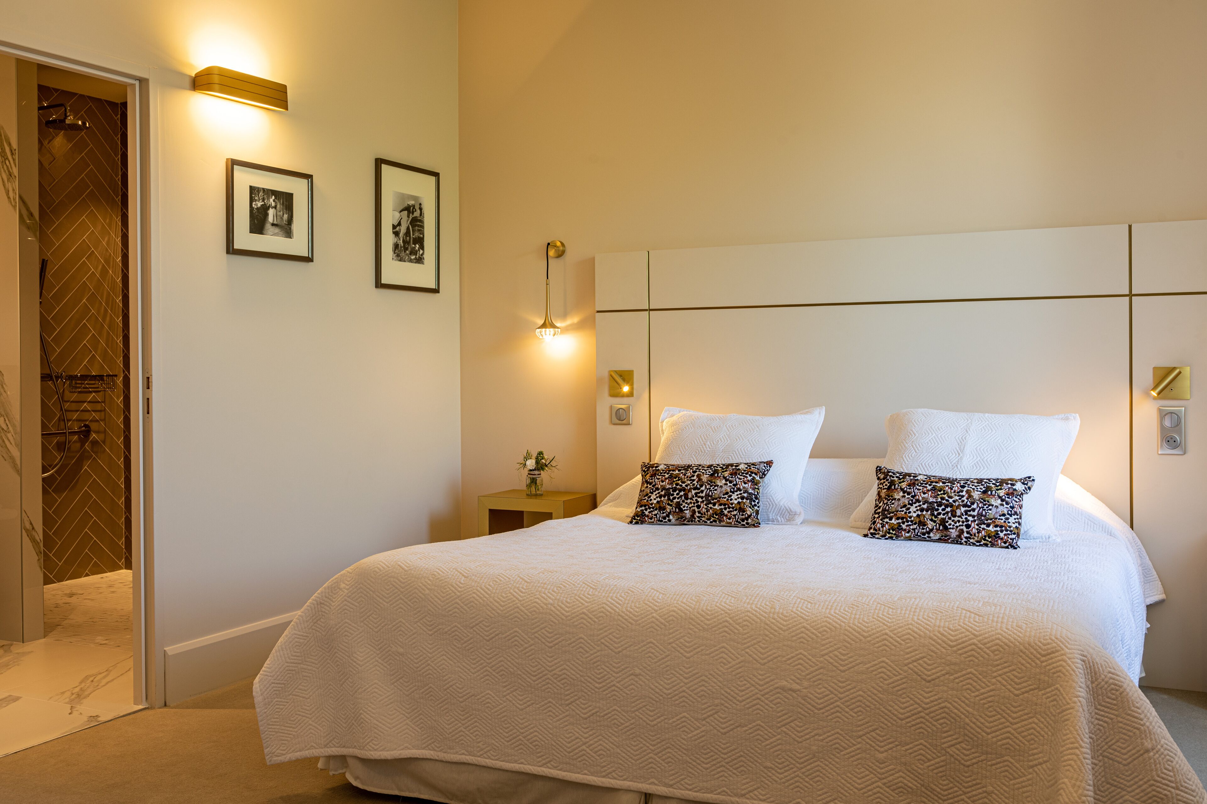 A neatly made bed with two pillows, a small bedside table with a lamp, and two framed pictures on the wall.