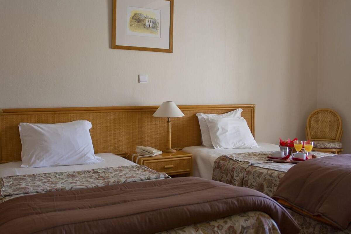 A hotel room with two beds, a bedside table with a lamp, a framed picture on the wall, and a small table with breakfast items.