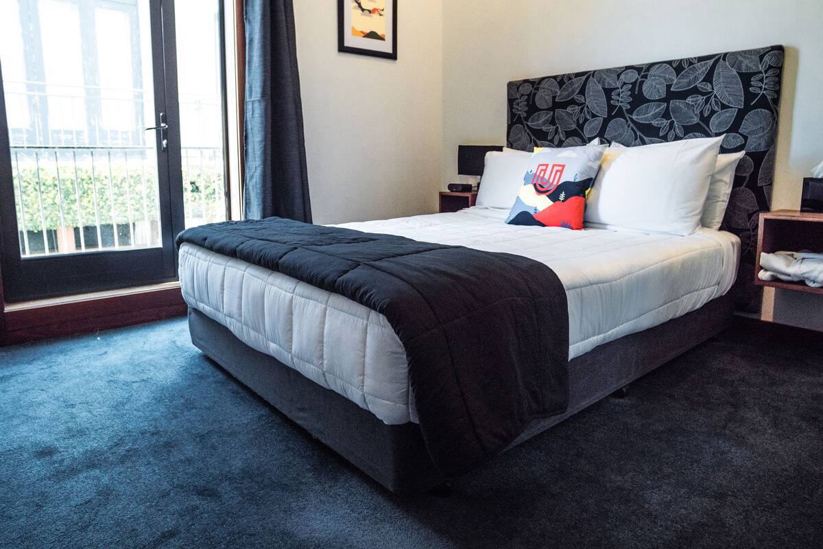 A hotel room with a large bed, a nightstand, a bedside table, a window with curtains, and a framed picture on the wall.
