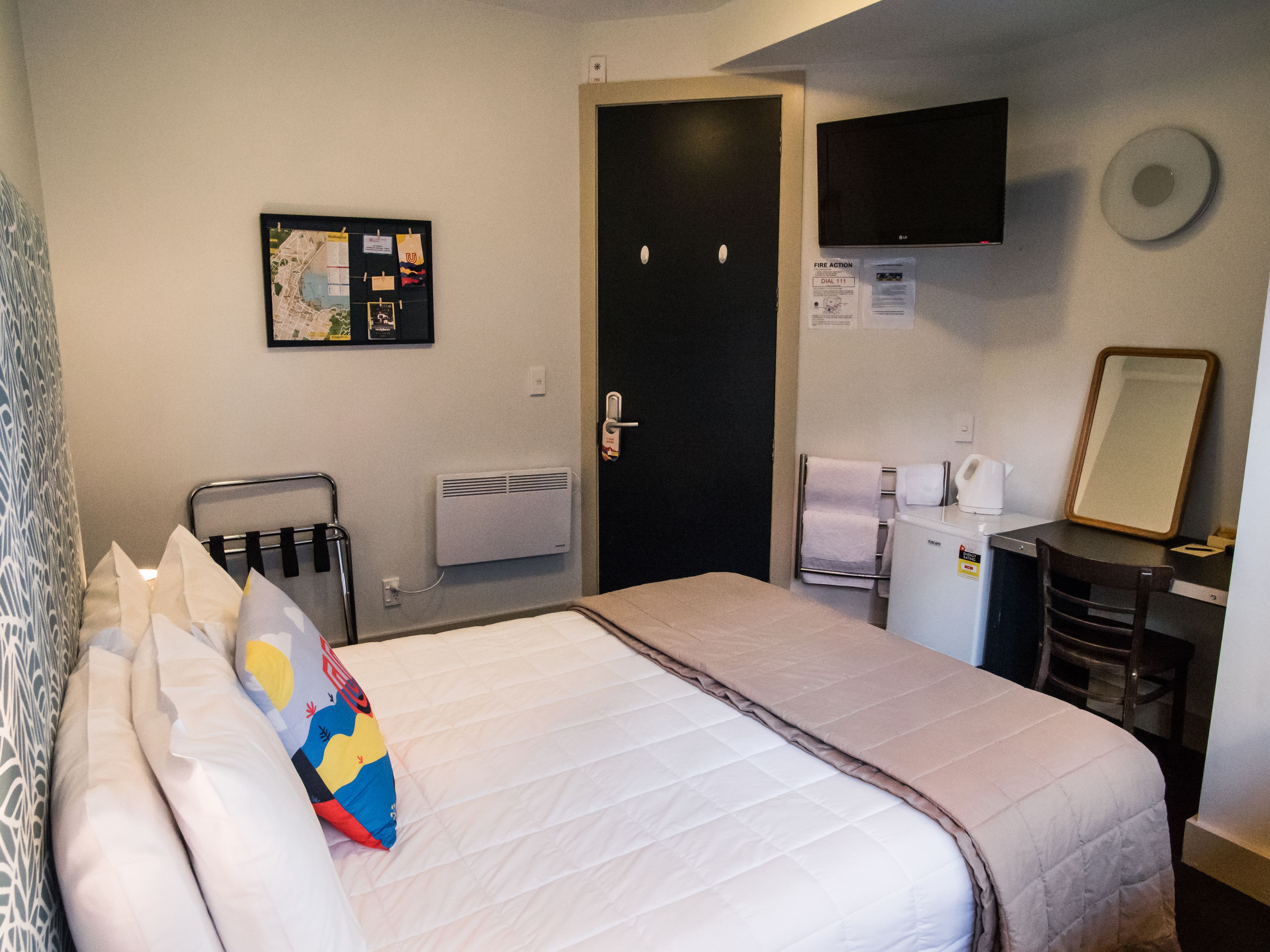 A hotel room with a bed, a desk, a chair, a small kitchenette, and a door.