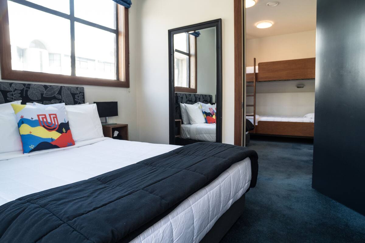 A hotel room with a large bed, a bedside table, a mirror, a window, and a bunk bed in the background.