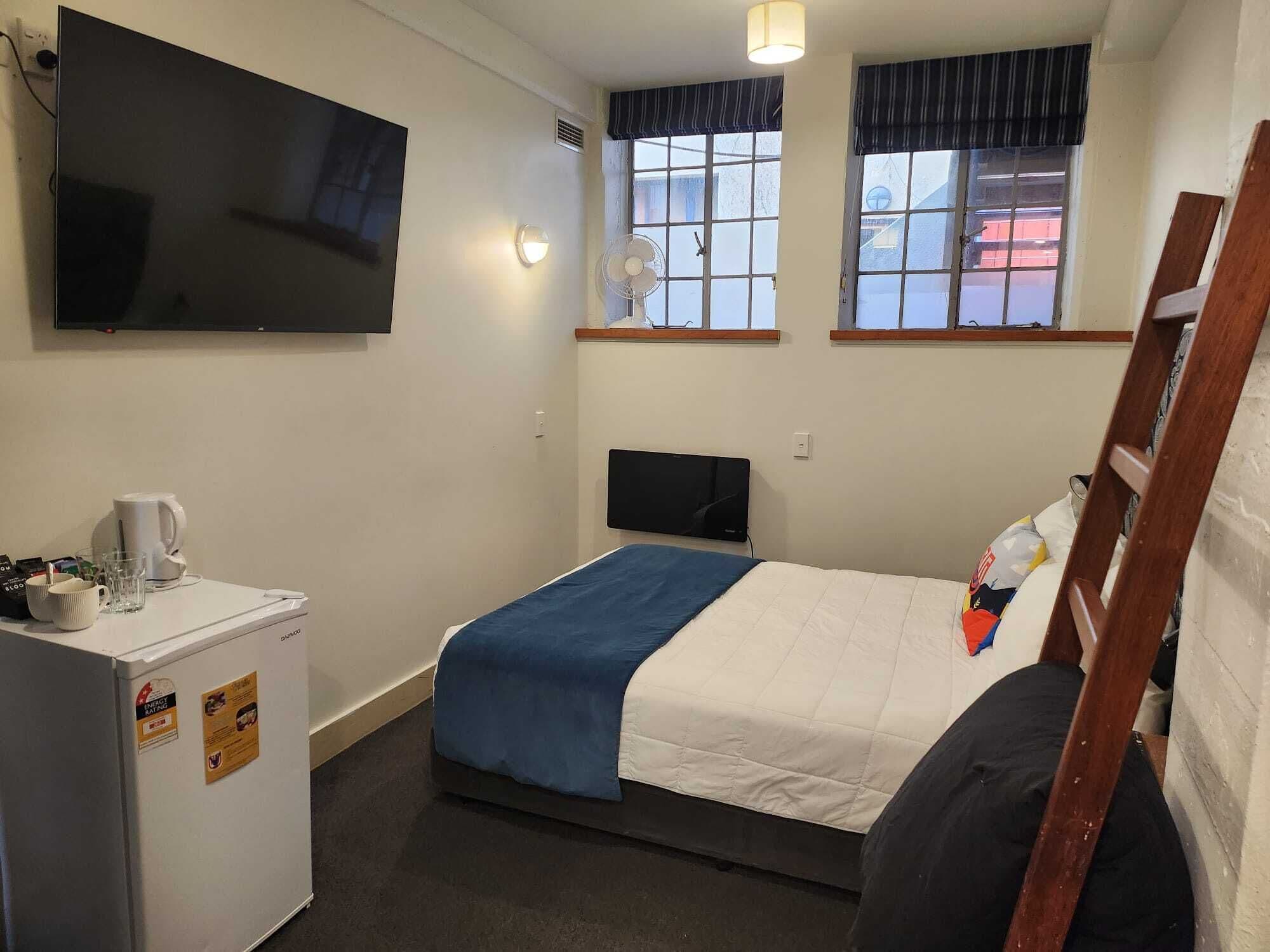 A small, simple room with a bed, a desk, a television, and a window.