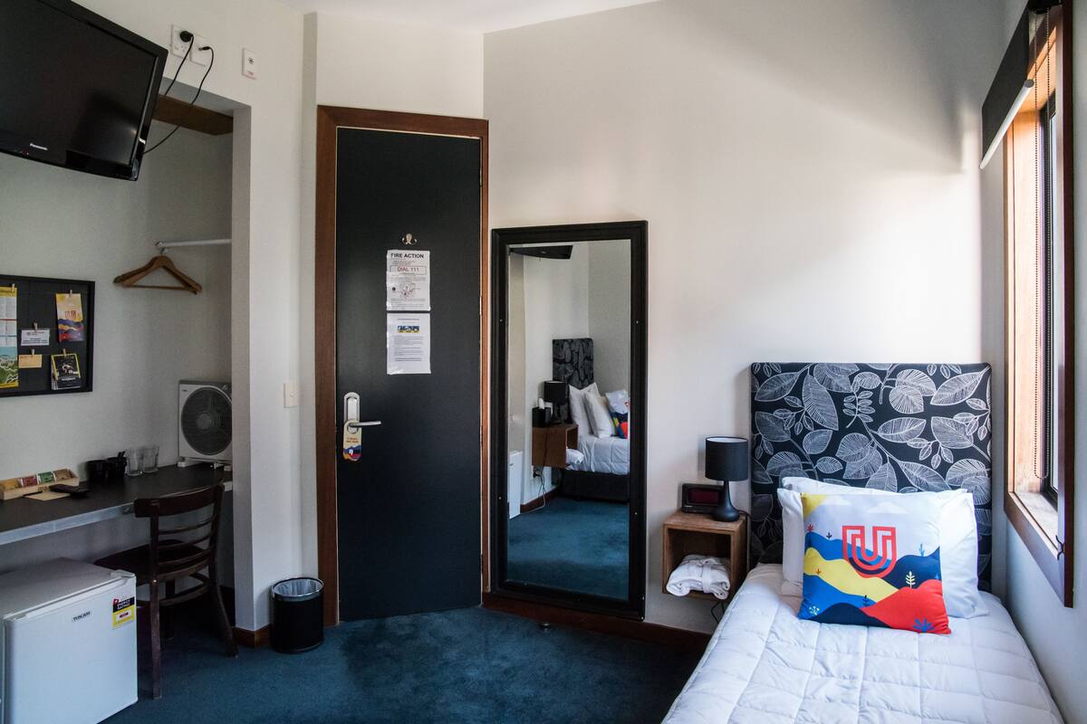 A hotel room with a bed, a desk with a computer, a chair, a small table with a lamp, a mirror, and a wall-mounted TV.