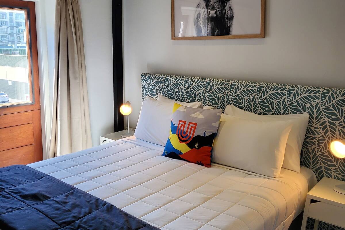 A neatly made bed with a white and blue quilt, a colorful pillow, and a framed artwork of a bull on the wall.