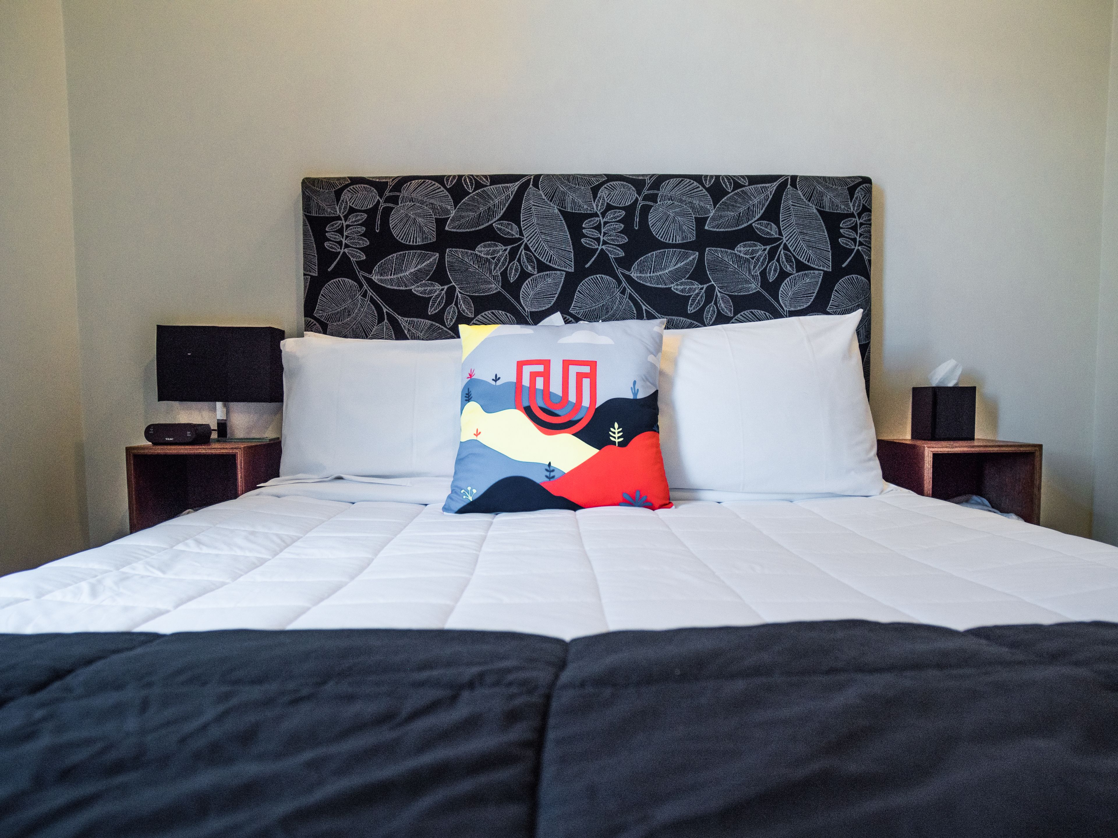 A neatly made bed with white linens and a black blanket, two pillows with a colorful design, a black and white patterned headboard, a bedside table with a lamp, and a tissue box on the nightstand.
