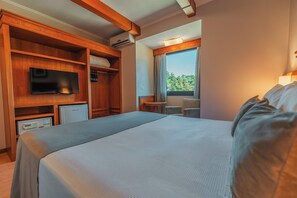 A hotel room with a large bed, a television, a minibar, and a window with a view of greenery. - Hotel Laghetto Premio (Gramado)