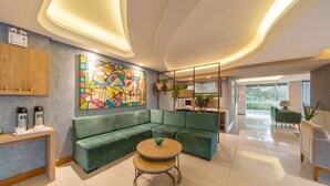 A modern hotel lobby with a green sectional sofa, a round wooden table, and a colorful abstract painting on the wall. - Hotel Laghetto Premio (Gramado)
