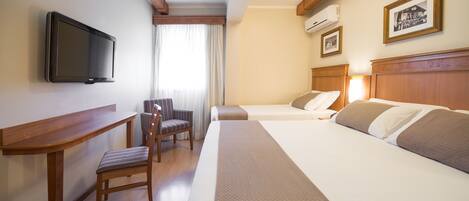 Una habitación de hotel con televisión montada en la pared, un escritorio, dos camas, una silla y una ventana con cortinas.