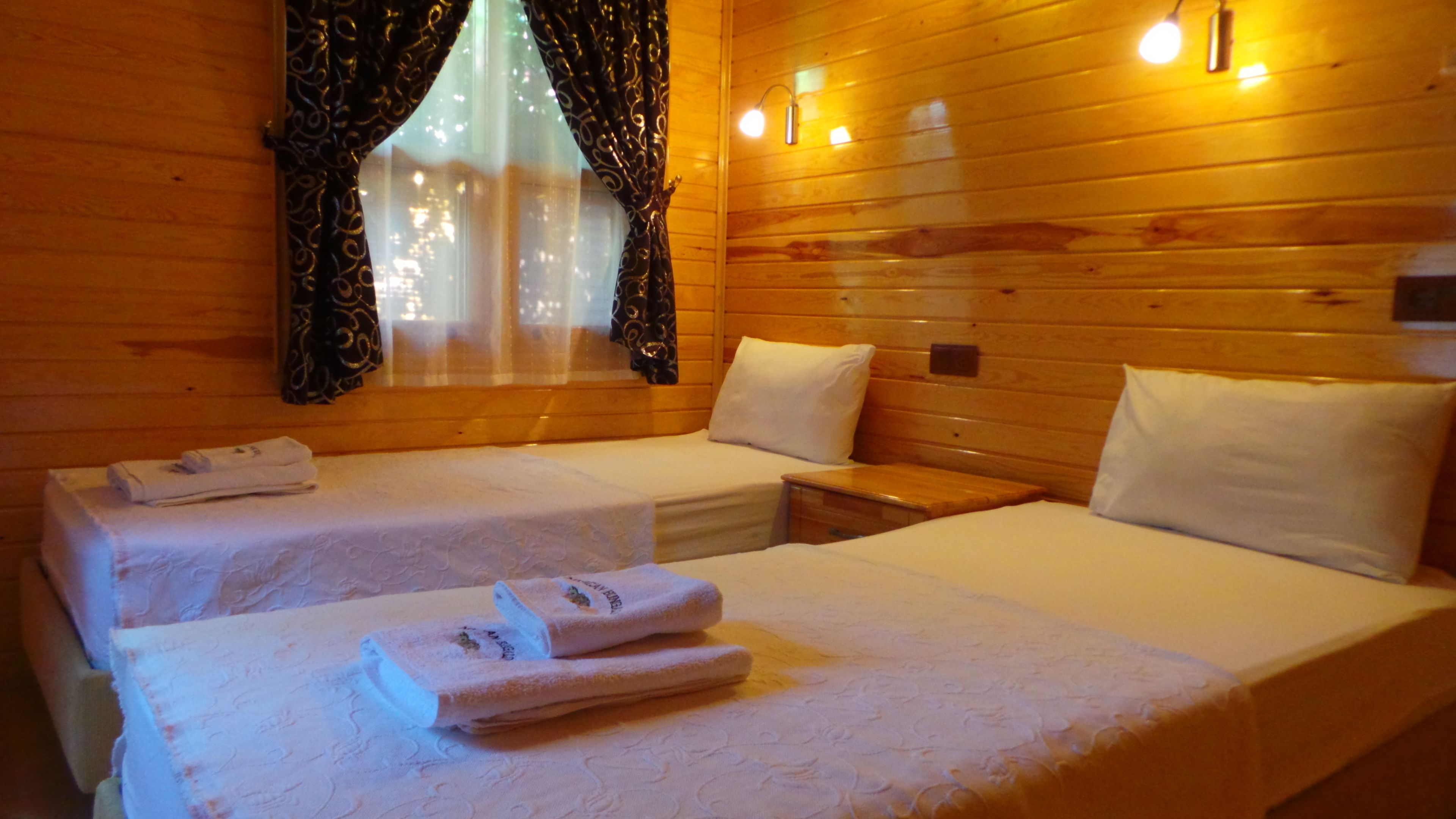A room with two beds, each with a white bedspread and pillows, a small wooden nightstand, and a window with curtains.