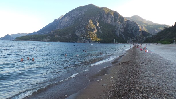 Una playa con gente nadando y tomando el sol, una montaña rocosa al fondo y un cielo despejado.