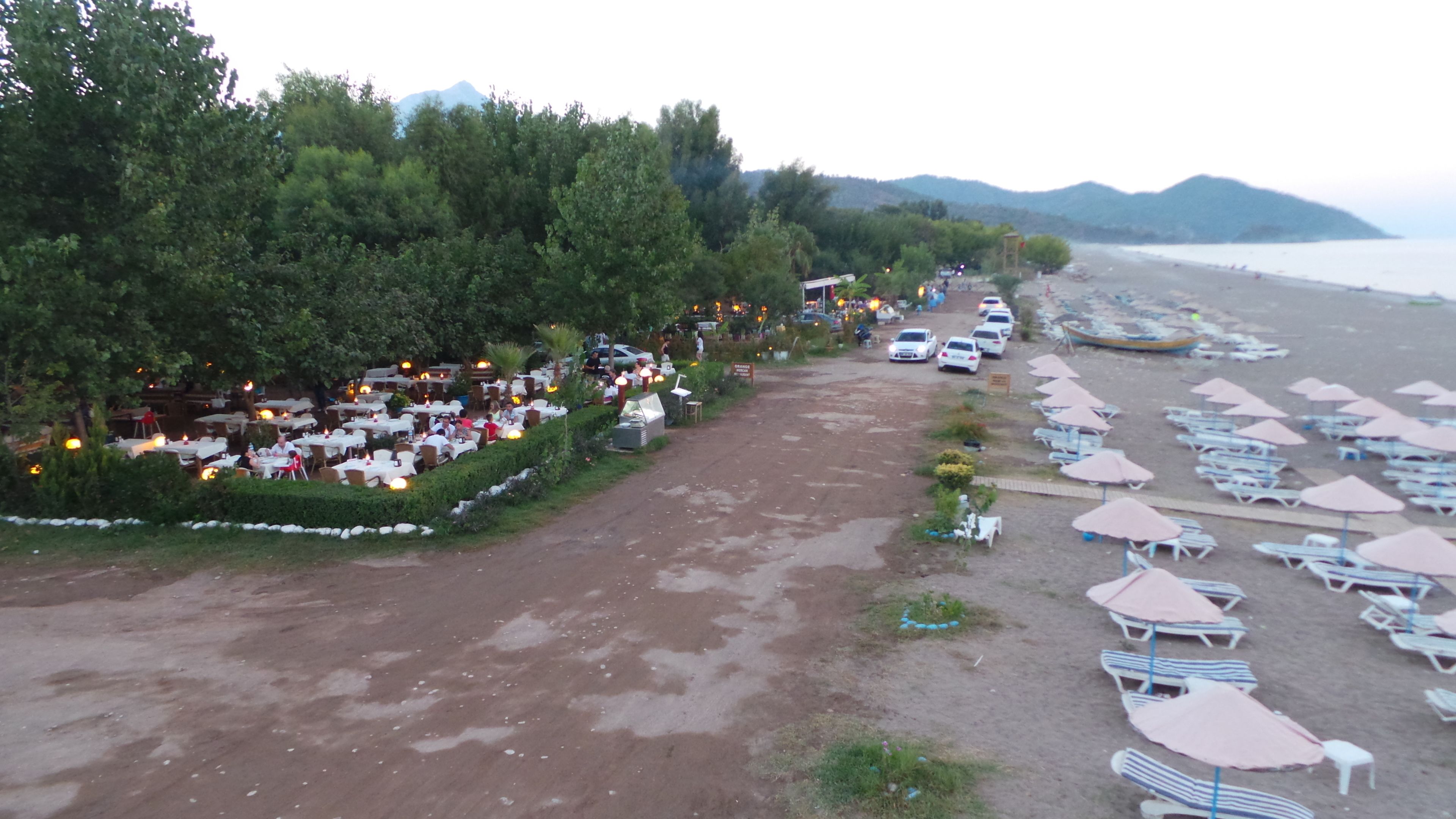 An outdoor restaurant with tables and chairs, a dirt path, and beach umbrellas.
