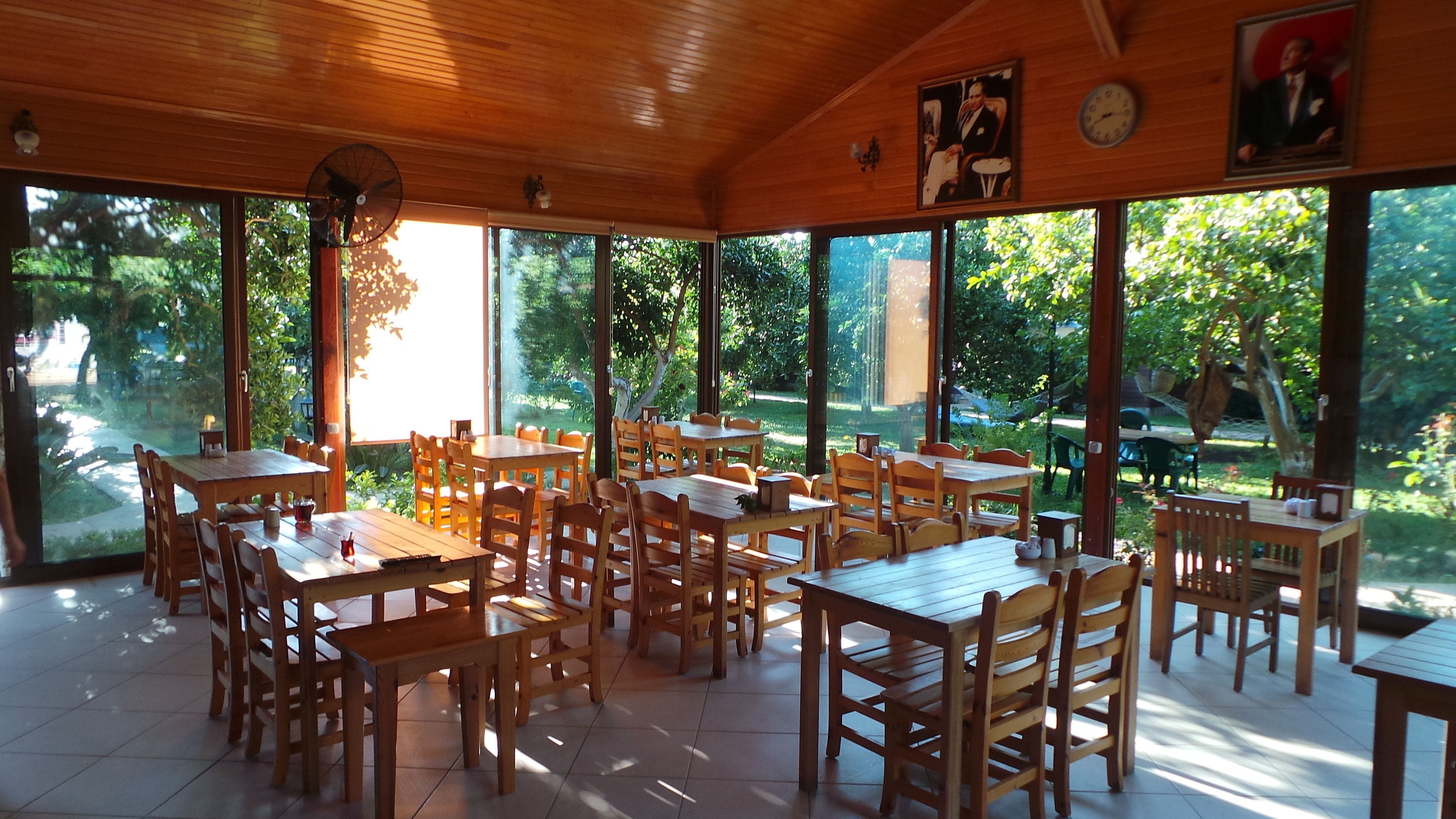 A restaurant with wooden tables and chairs, large windows, and a view of greenery.