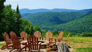 Un grupo de sillas Adirondack dispuestas alrededor de un brasero de piedra, con una vista panorámica de un valle boscoso y montañas al fondo.