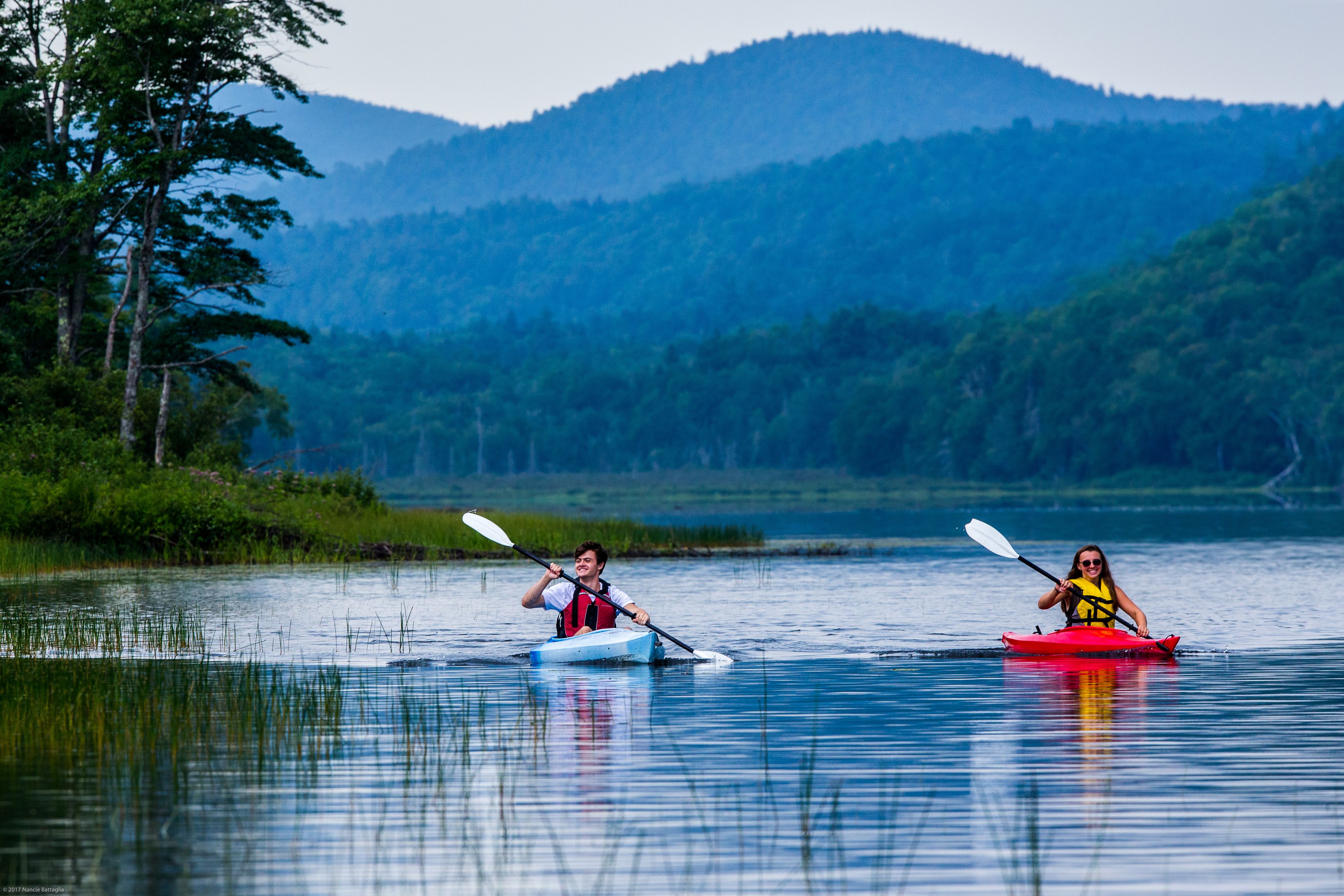 Two kayakers on a calm lake with forested hills in the background.