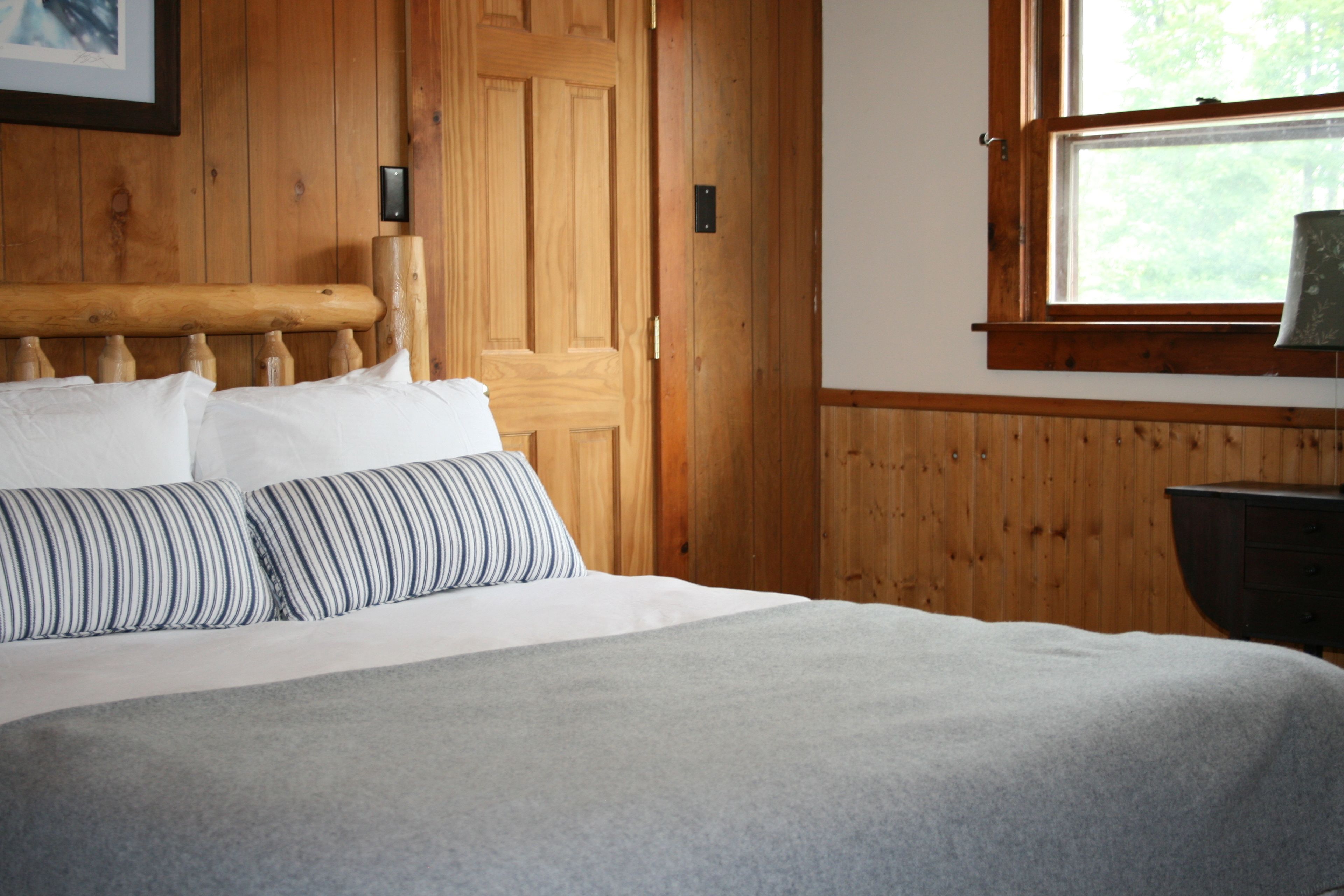 A bed with white bedding and striped pillows, a wooden headboard, and a window with a view of trees.
