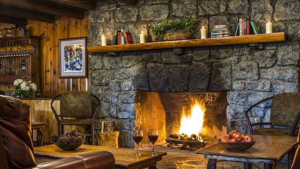 Una chimenea acogedora con un muro de piedra, un estante de madera con libros y velas, y una mesa con un cuenco de manzanas y copas de vino.