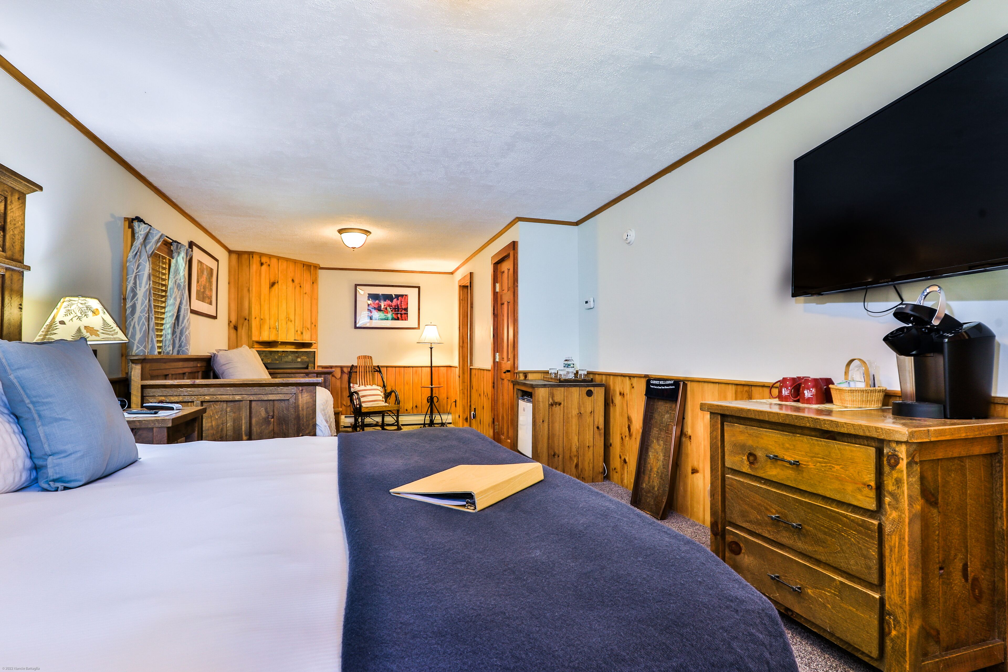 A room with a bed, wooden dresser, TV, and a desk with a lamp.