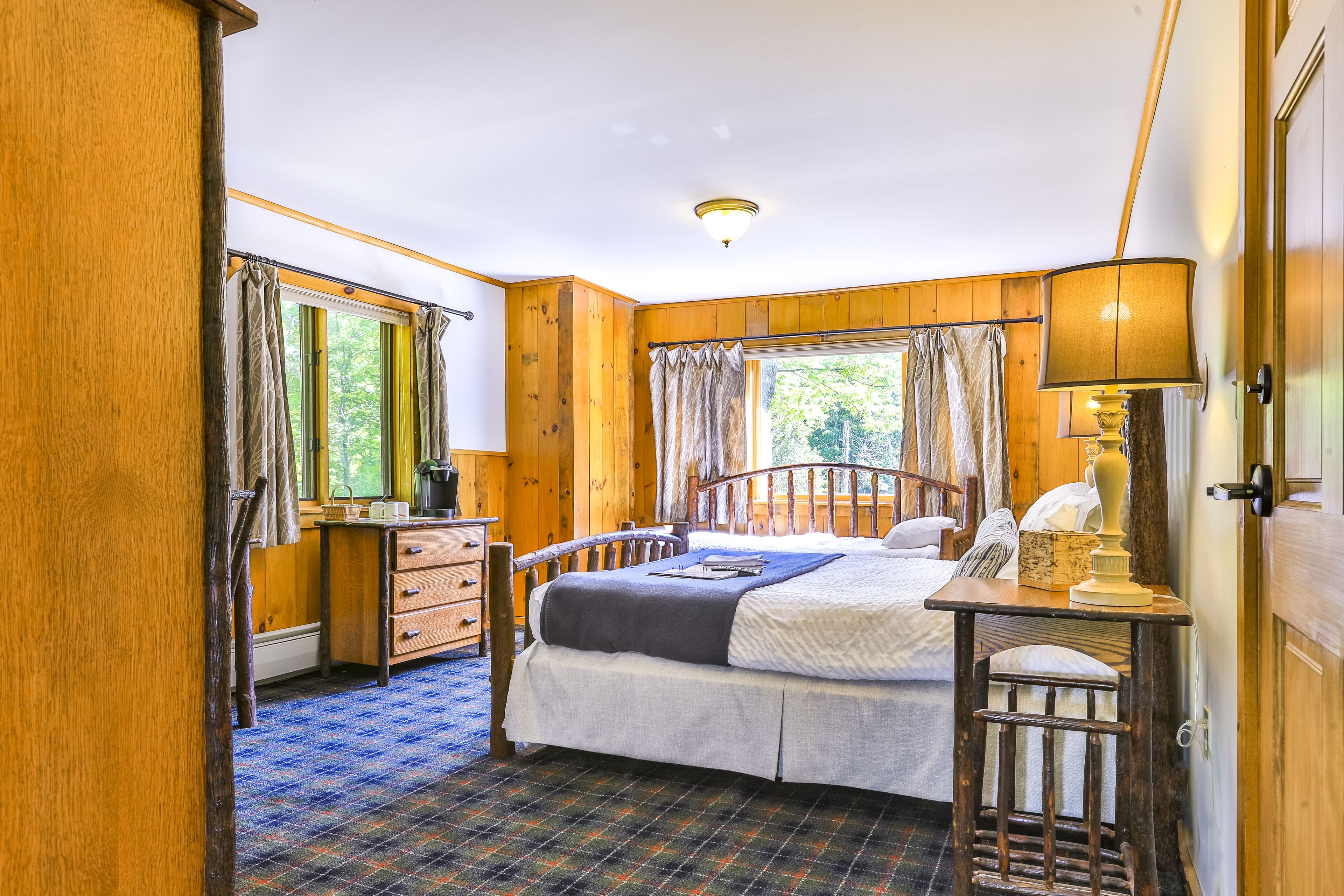 A wooden cabin bedroom with a bed, dresser, and a small table with a lamp.