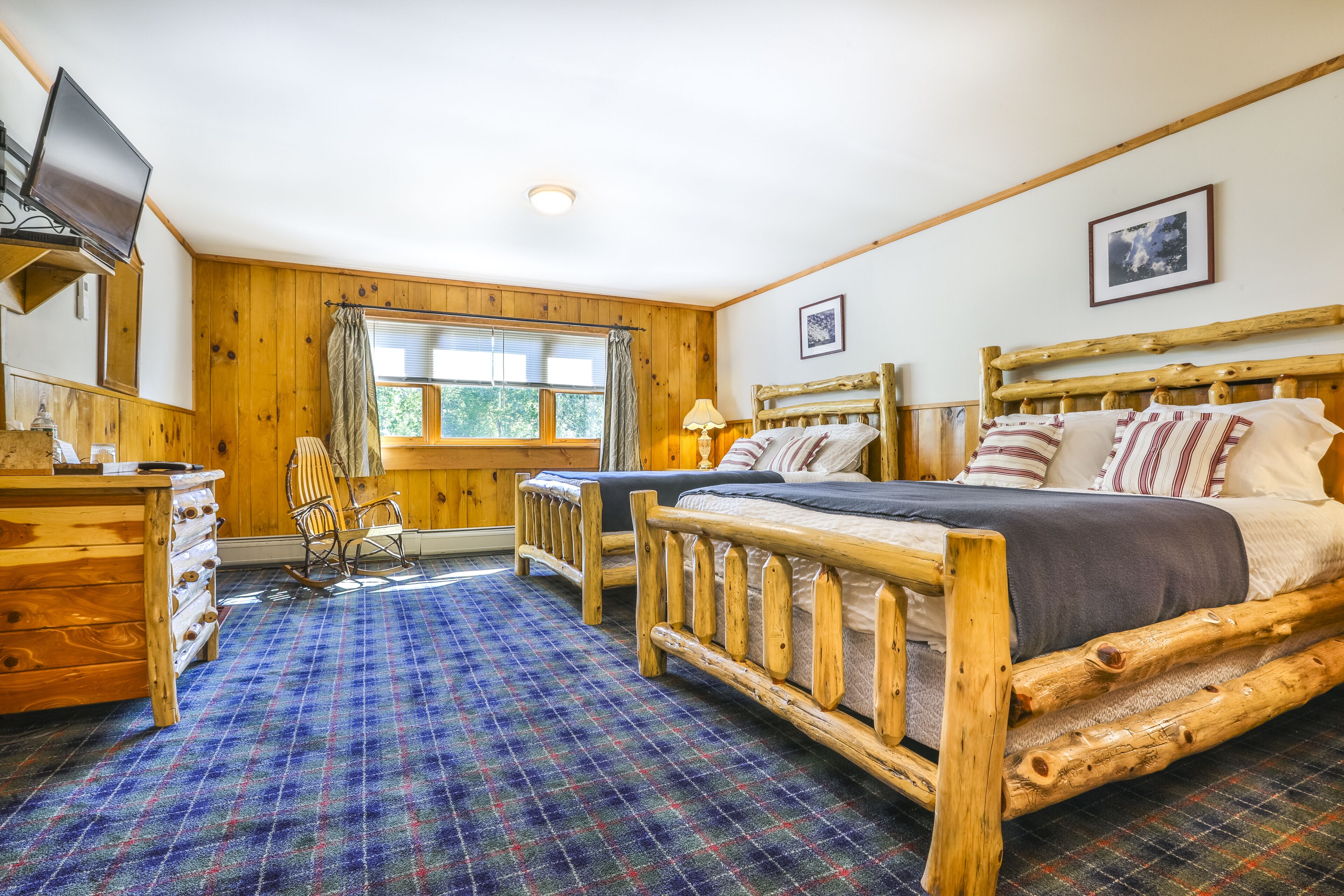 A wooden cabin-style room with two beds, a television, a dresser, and a plaid carpet.