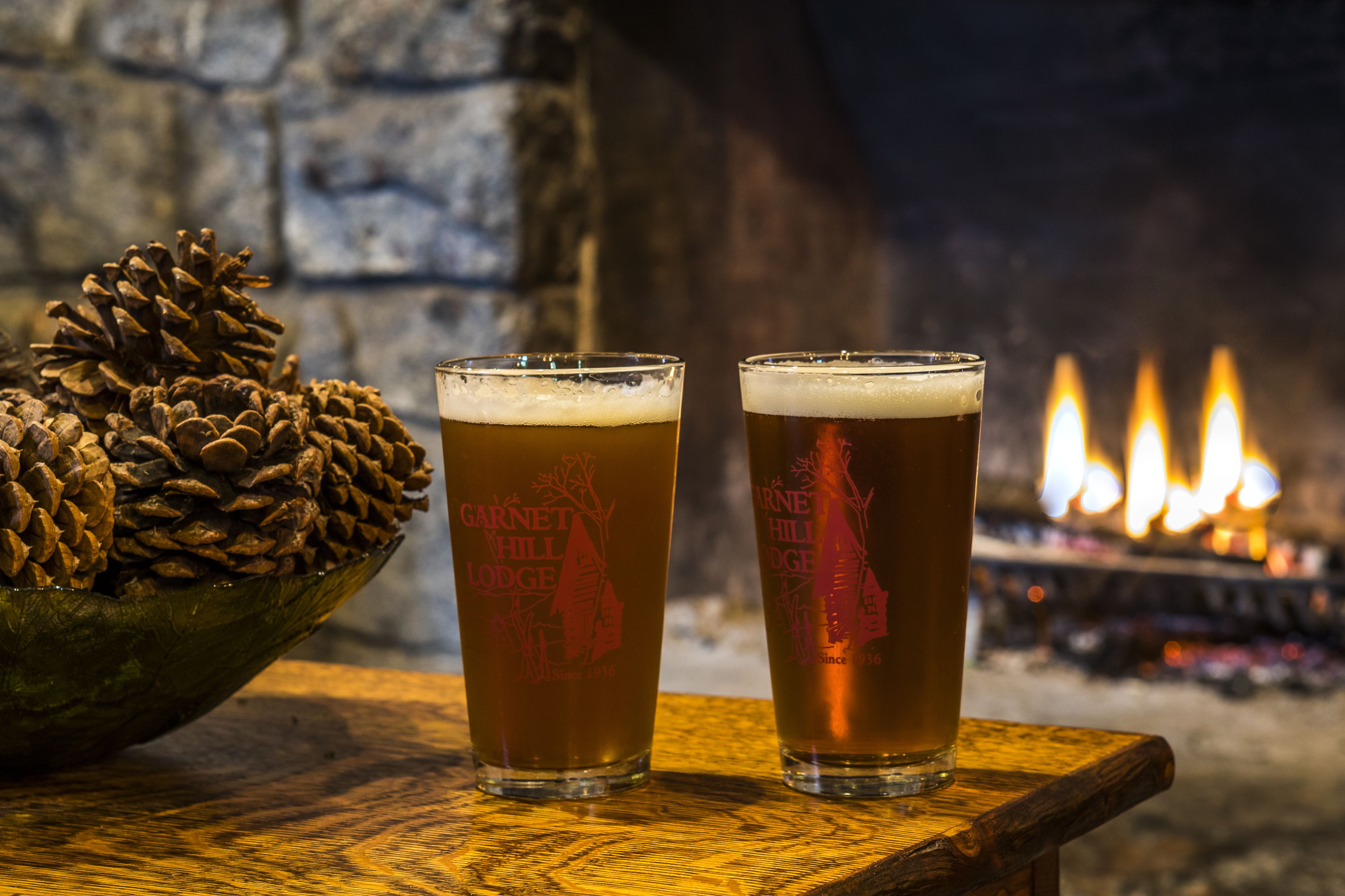 Two glasses of beer on a wooden table with a pine cone and a fireplace in the background.