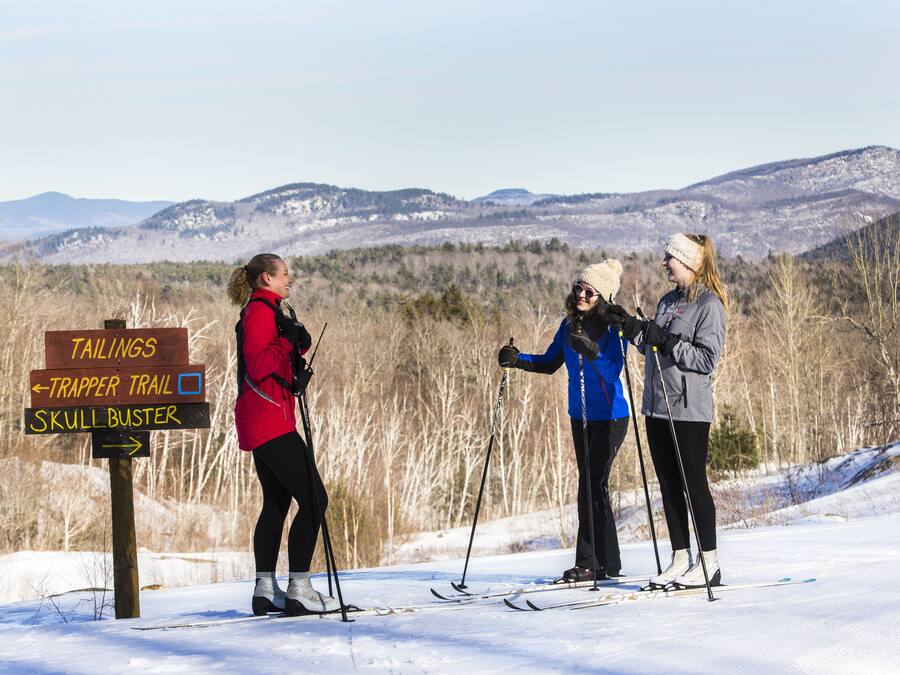 Three people standing on a snowy path with a signpost indicating directions to Tailings, Trapper Trail, and Skull Buster.