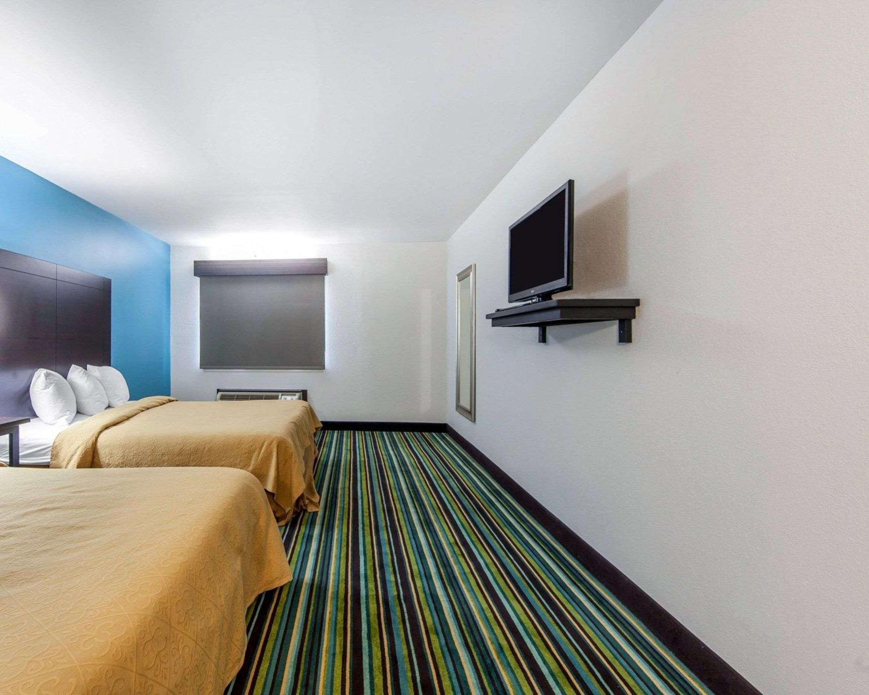 Habitación de hotel con dos camas, un televisor sobre un mueble bajo, una ventana con persianas y una alfombra a rayas.