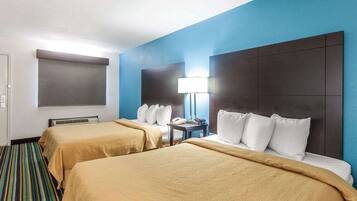 A hotel room with two beds, a large window with blinds, a bedside table with a lamp, and a striped carpet.