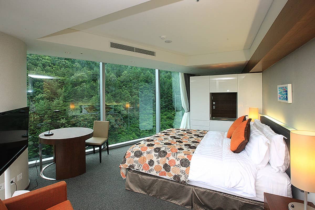 A hotel room with a large bed, a desk with a TV, a chair, and a view of a forest through large windows.