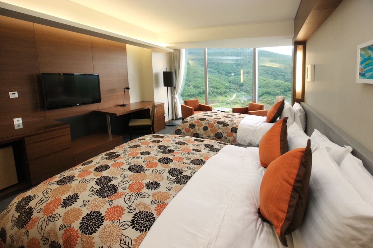 A hotel room with two beds, a TV, a desk, and a view of a mountainous landscape.