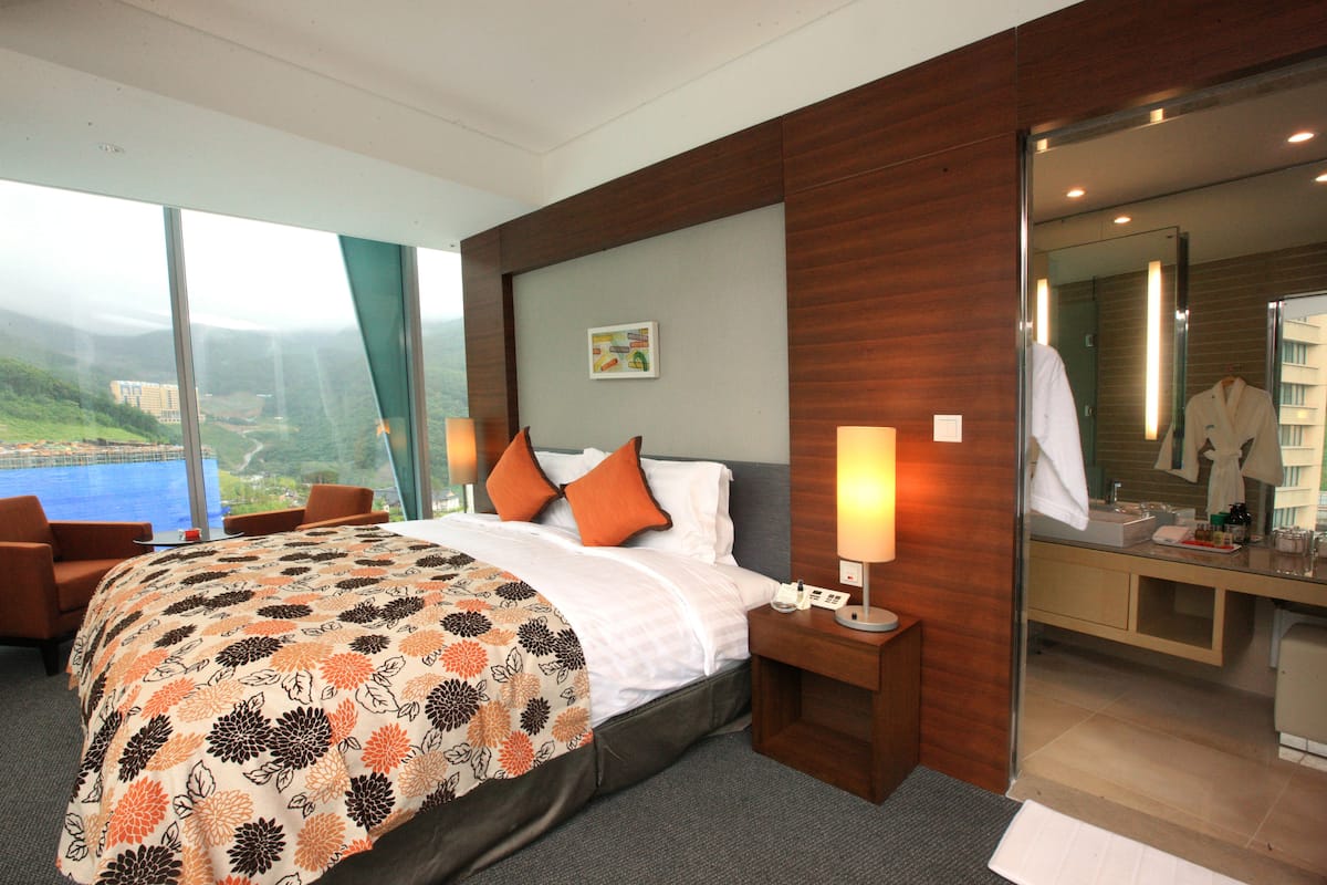 A hotel room with a large bed, two nightstands, a bathroom with a glass partition, and a view of a body of water.