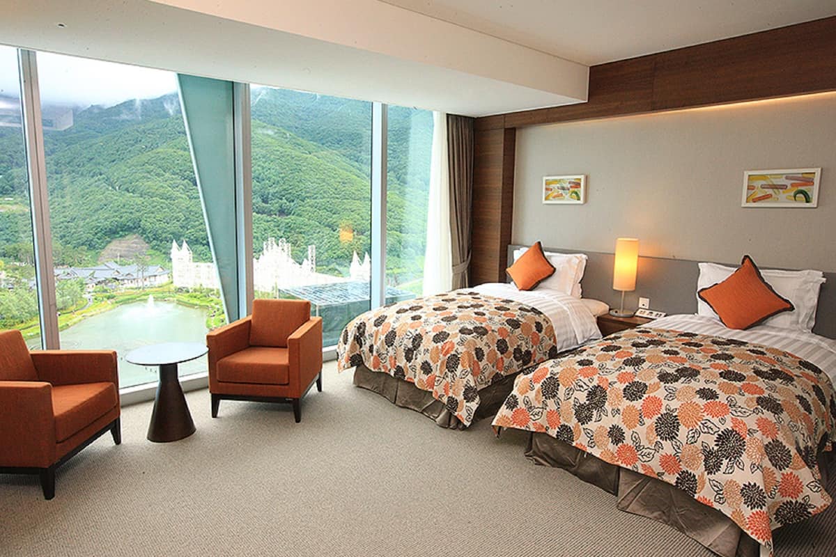 A hotel room with two beds, a small table, two armchairs, and large windows offering a view of a castle and greenery.