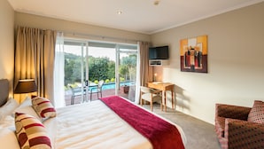 A hotel room with a large bed, a desk, a TV, and a view of a pool and greenery. - Decks of Paihia (Paihia)