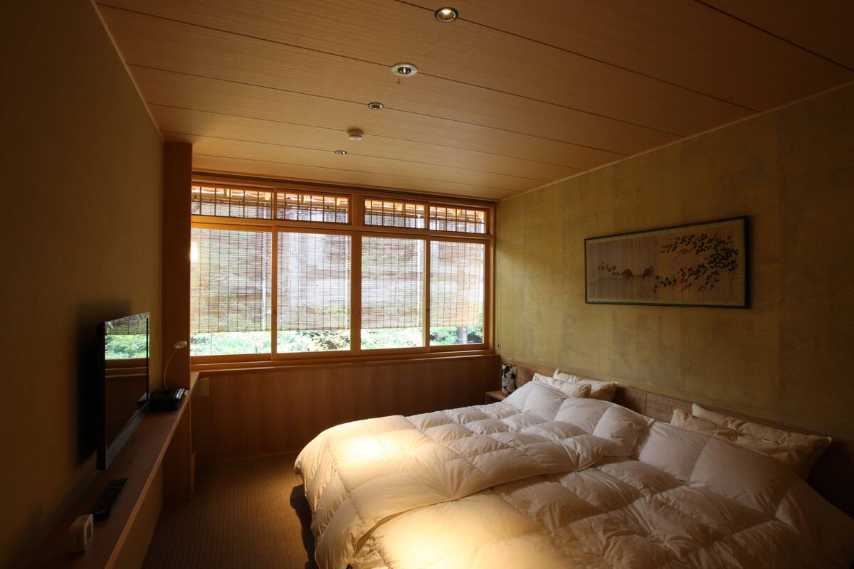 Une chambre dotée d’un grand lit, d’un bureau avec un ordinateur et d’une fenêtre avec des stores.