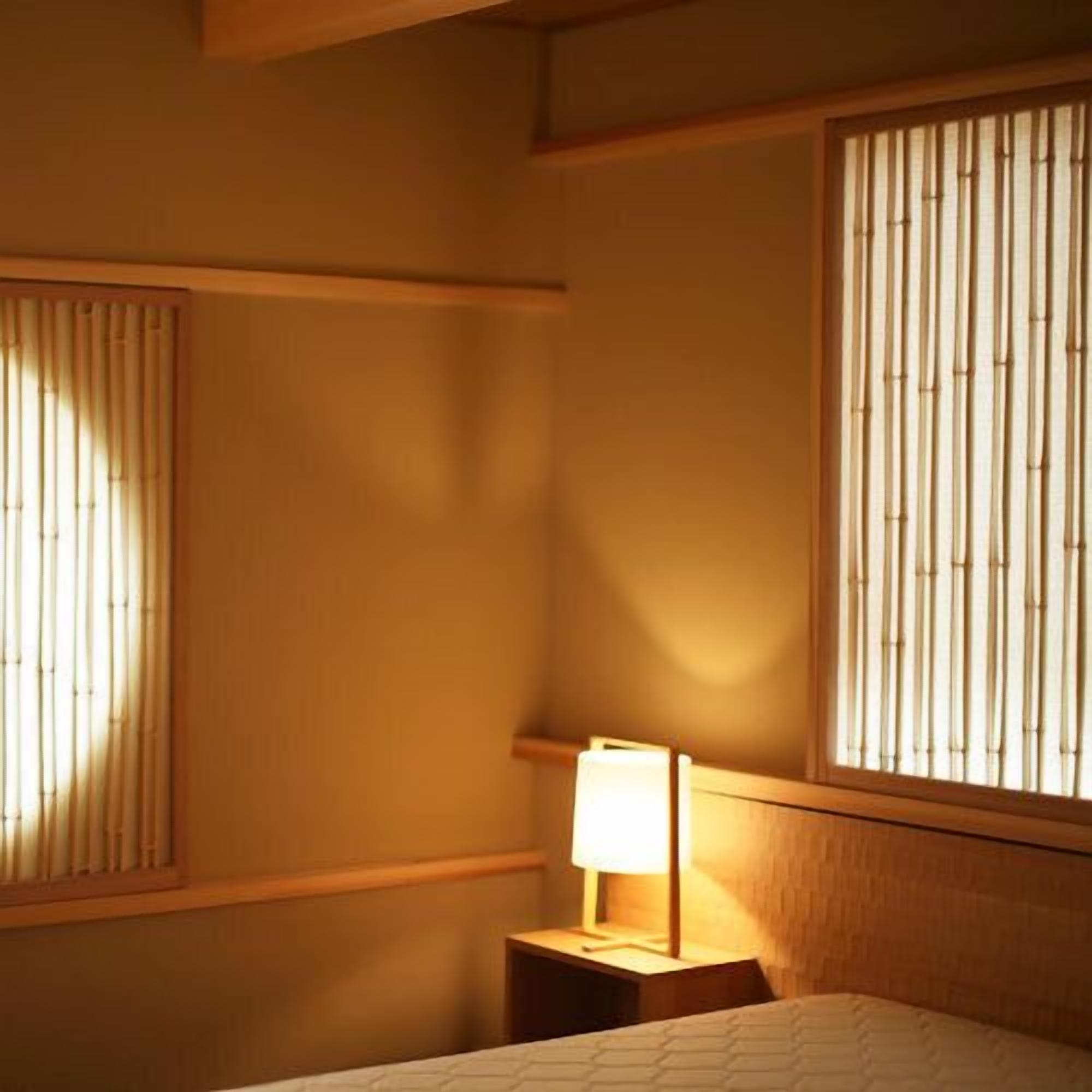 Hotellihuone, jossa on sänky, yöpöytä lampun kanssa ja ikkuna, jossa on pystysuorat sälekaihtimet.