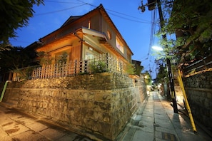 A two-story house with a stone wall and a wooden fence, located on a narrow street with a paved sidewalk.