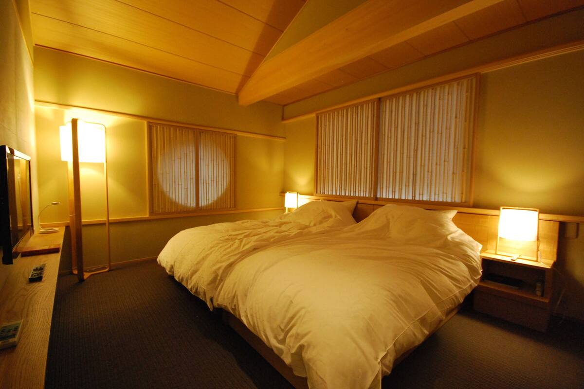 Un grand lit avec du linge de lit blanc, deux tables de chevet avec des lampes, une tête de lit en bois et une grande fenêtre avec des rideaux.