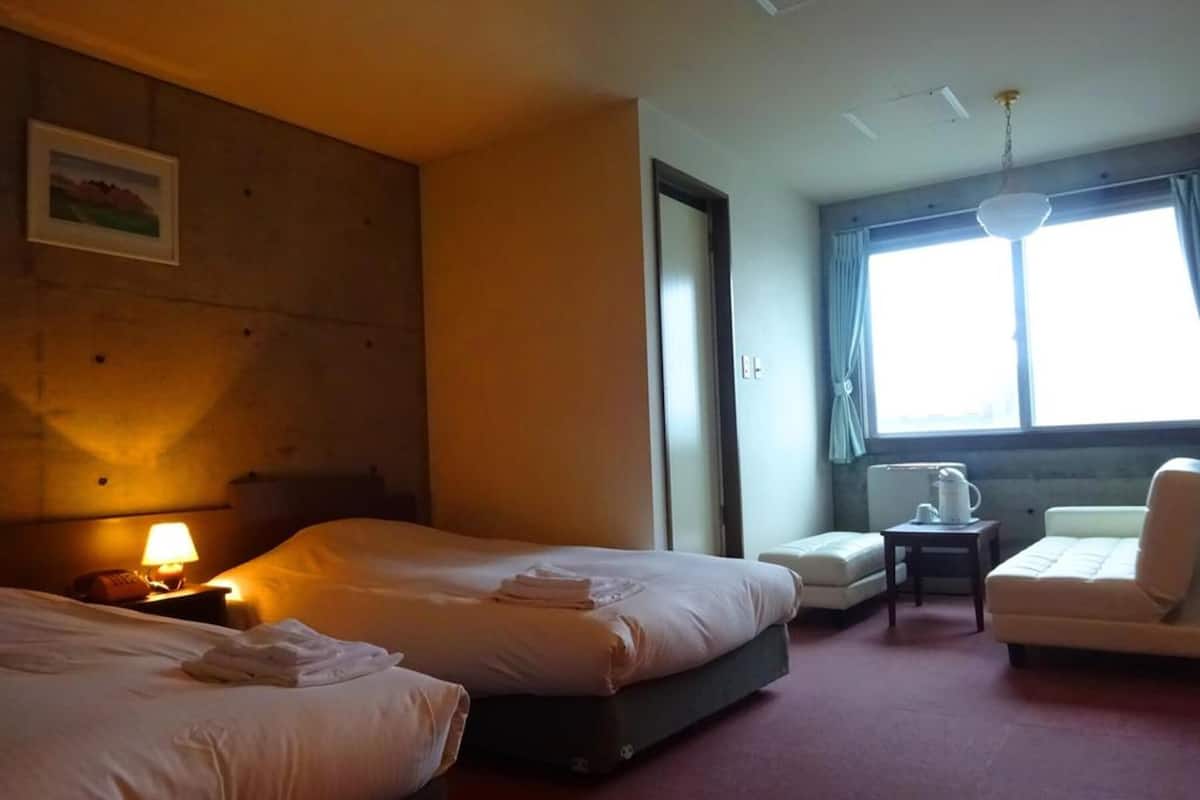 A hotel room with two beds, a small table with a lamp, a chair, and a window with curtains.