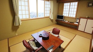 A traditional Japanese room with tatami flooring, a wooden table with a remote control, a television on a shelf, and a view of a cityscape through large windows. - Hakodate Motomachi Hotel (Hakodate)