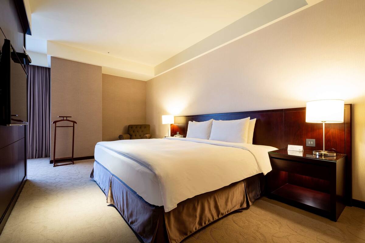 A hotel room with a large bed, bedside lamps, a wooden nightstand, a wardrobe, and a desk with a lamp.