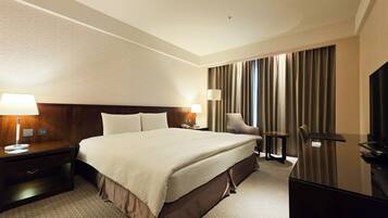 Een hotelkamer met een groot bed, nachtkastjes met lampen, een bureau met een stoel, een televisie en een groot raam met gordijnen.