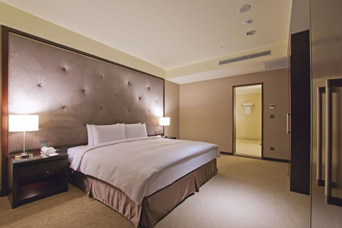 A hotel room with a large bed, two bedside tables with lamps, a wardrobe, and a bathroom visible through an open door.